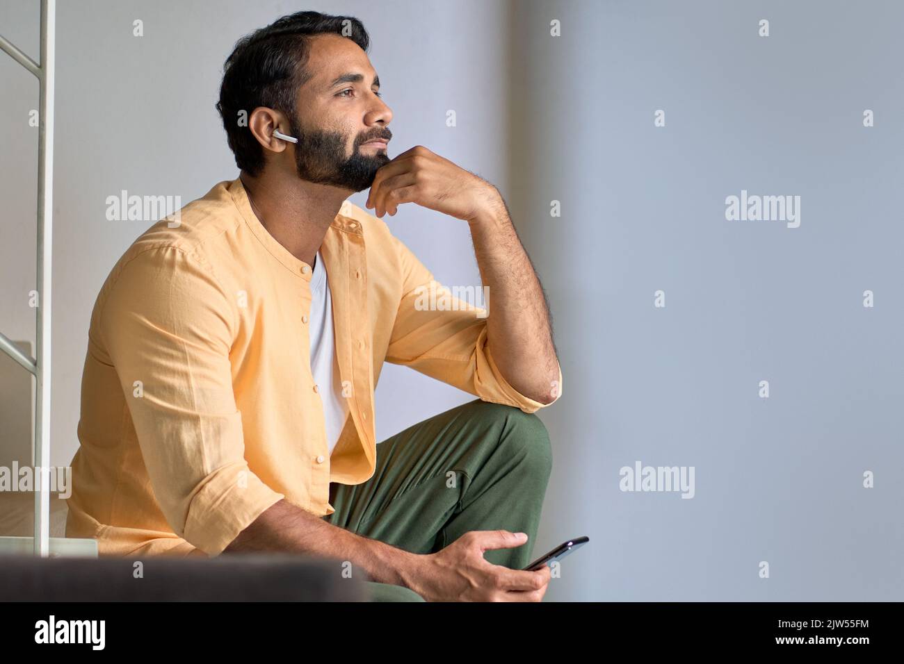 Indischer Mann, der zu Hause mit Ohrstöpseln sitzt und Podcast über das Mobiltelefon hört. Stockfoto