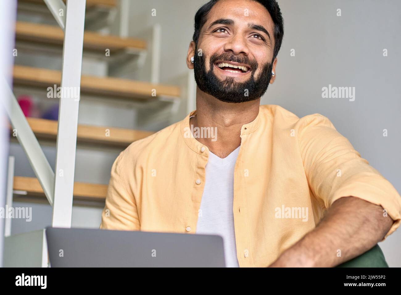Glücklich lachende ethnische indische Geschäftsmann trägt Ohrstöpsel mit Laptop. Stockfoto