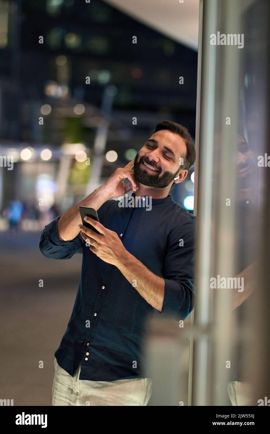 Ein indischer Mann mit Ohrstöpseln hält das Telefon und hört nachts mobile Musik. Stockfoto
