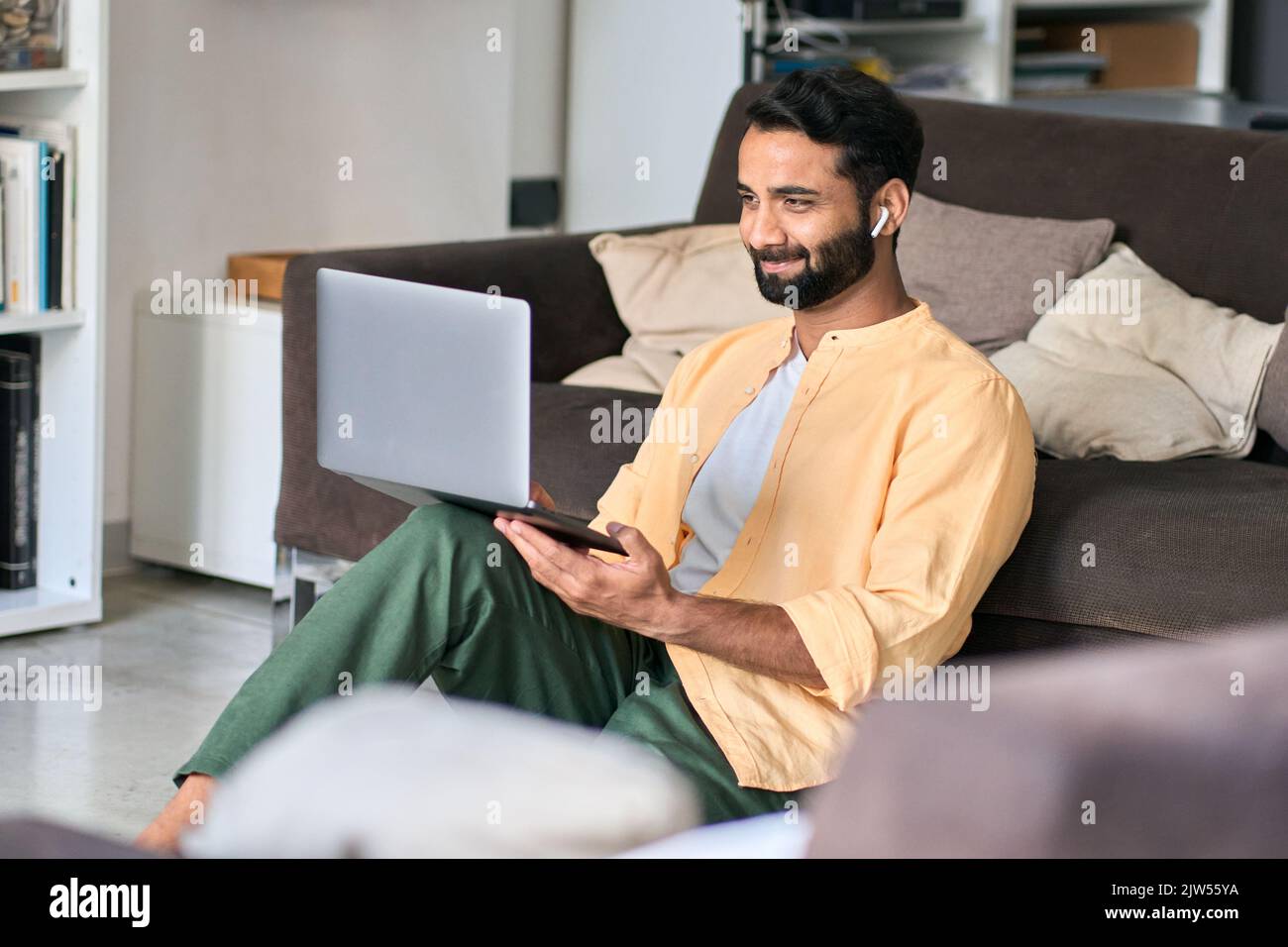 Glücklicher indischer Mann, der zu Hause mit einem Ohrstöpsel und einem Laptop sitzt. Stockfoto