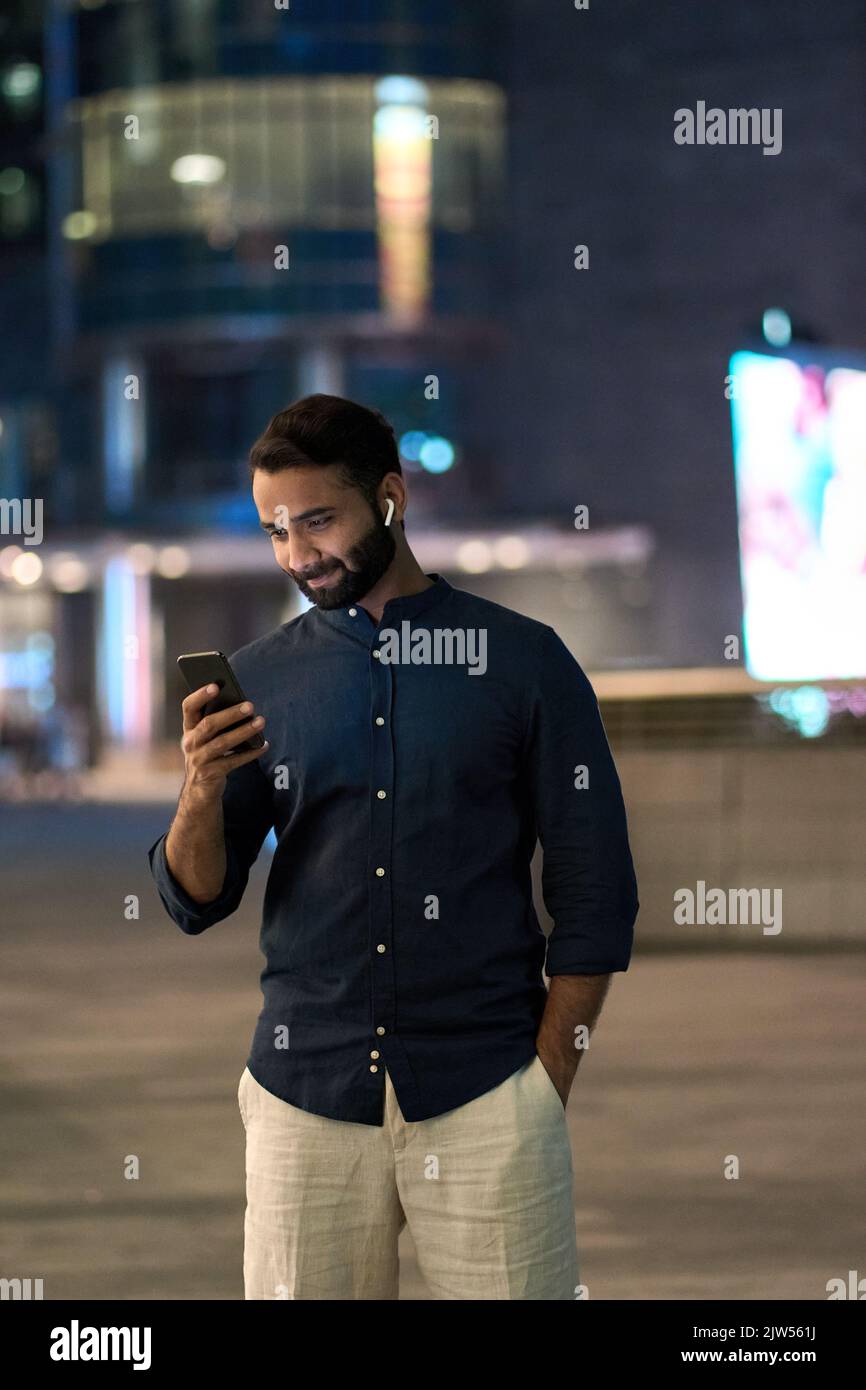 Indischer Mann mit Ohrstöpseln, der auf der nächtlichen Stadtstraße mit einem Mobiltelefon stand. Stockfoto