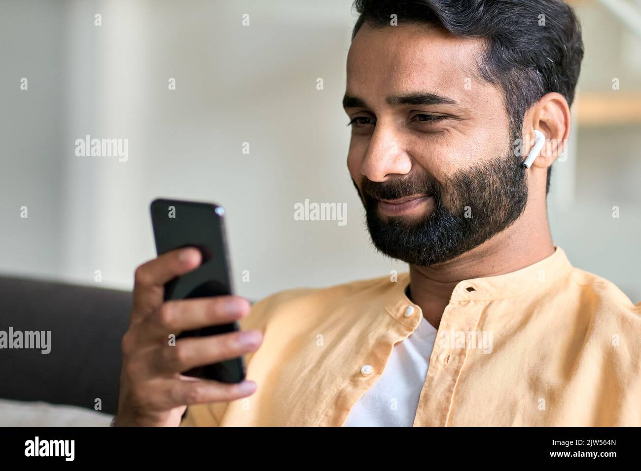 Lächelnder indischer Mann, der zu Hause mit Kopfhörern per Smartphone sitzt. Stockfoto