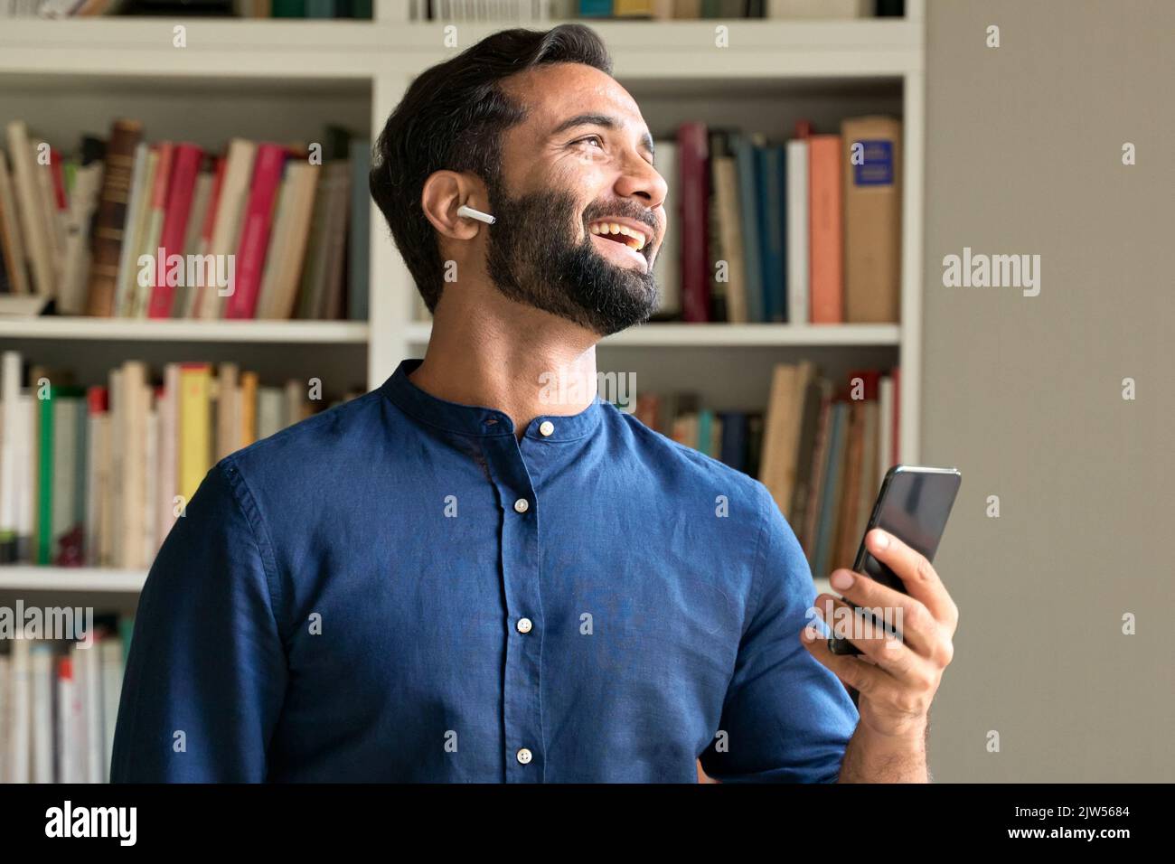 Glücklicher indischer Geschäftsmann bei der Arbeit, der Ohrstöpsel mit dem Smartphone trägt. Stockfoto