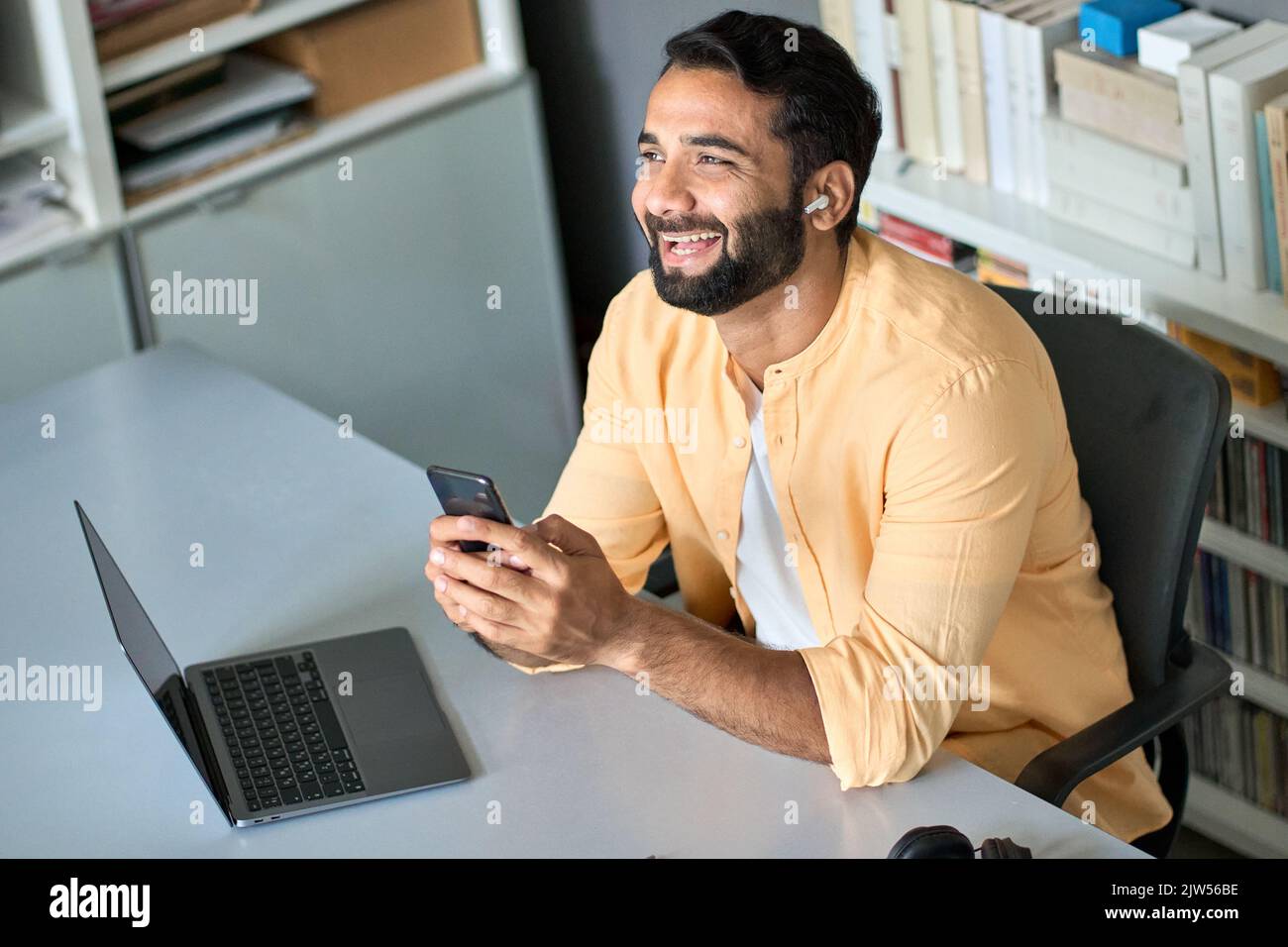 Glücklicher indischer Geschäftsmann bei der Arbeit, der Ohrstöpsel mit dem Handy trägt. Stockfoto