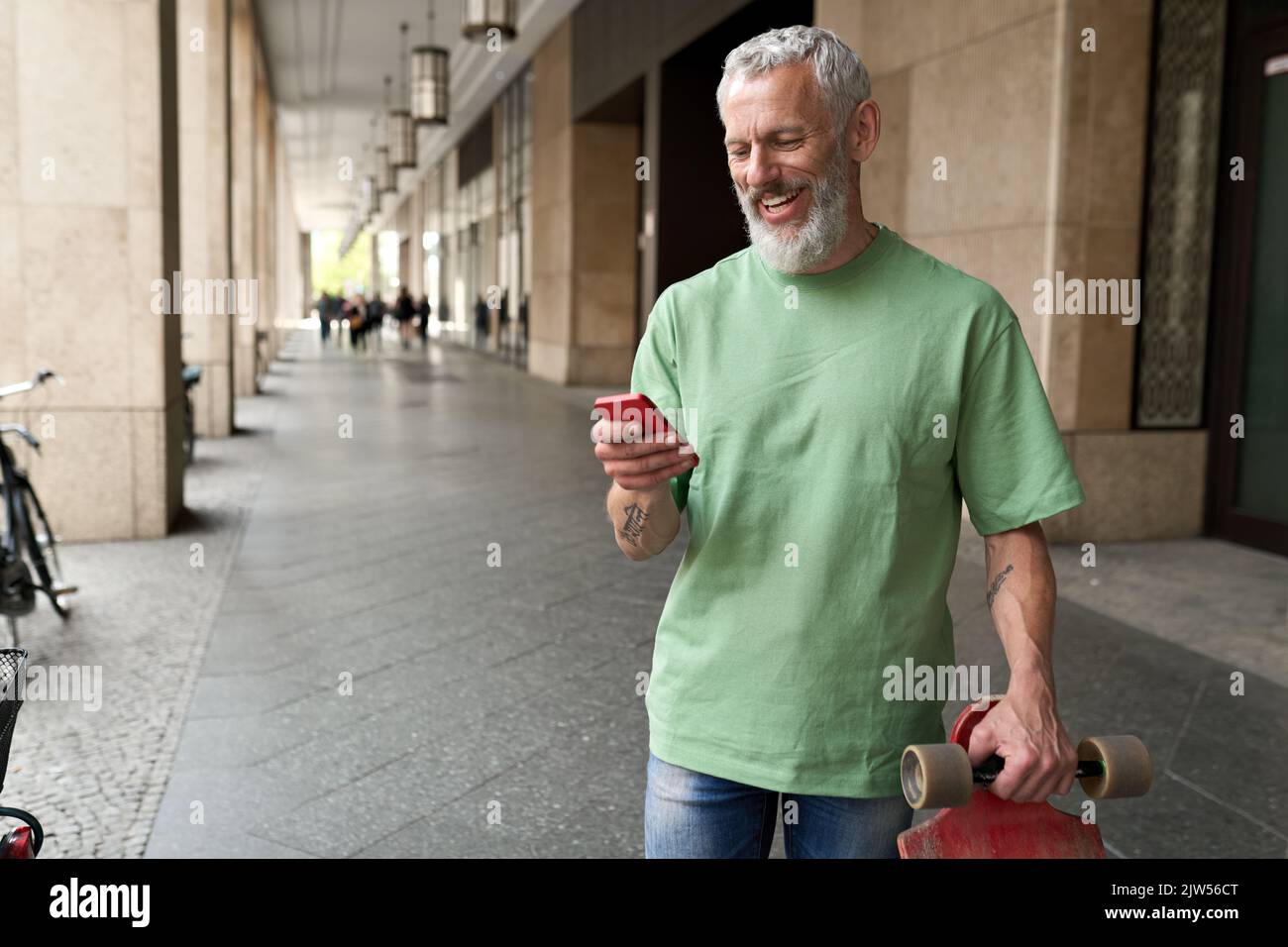 Lächelnder alter, reifer Hipster-Skater mit Handy, der in der Stadt steht. Stockfoto