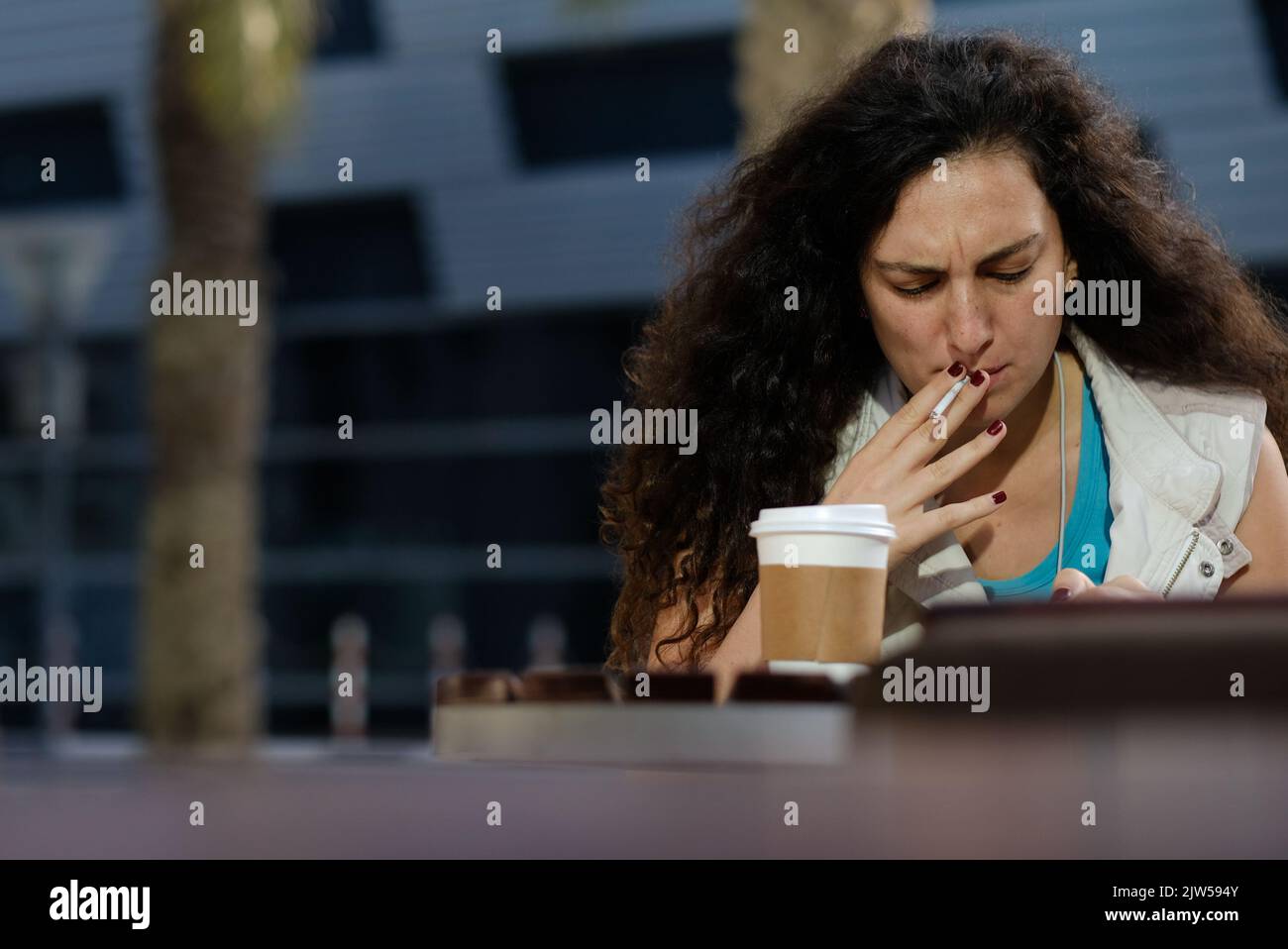 Frau mit langen lockigen Haaren und kastanienbraunen Nägeln macht mittags eine Zigarette- und Kaffeepause vor einem Café. Frau schaut mit einem Stirnrunzeln auf das Telefon. Stockfoto