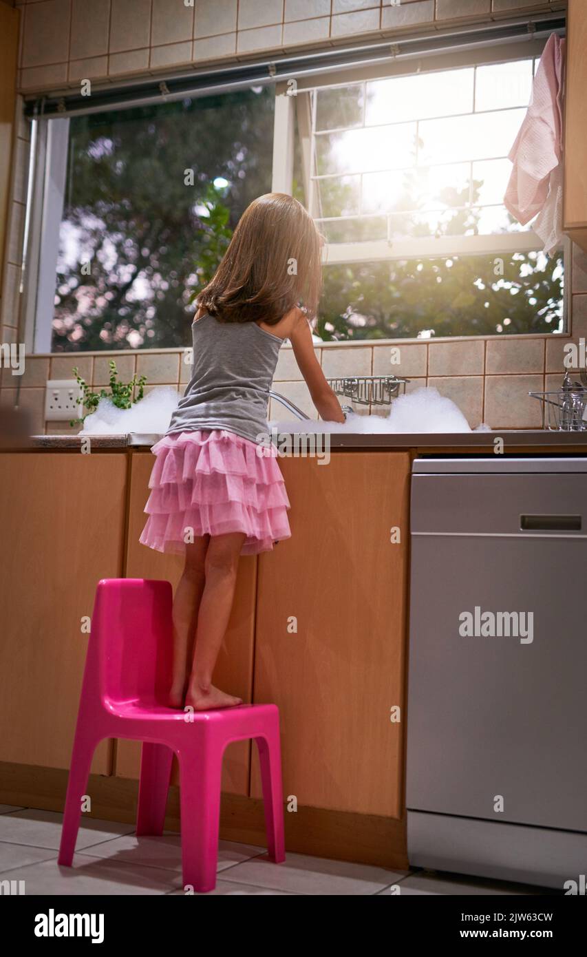 Ein junges Mädchen, das auf einem Stuhl steht, um das Geschirr an einem Spülbecken zu waschen. Stockfoto