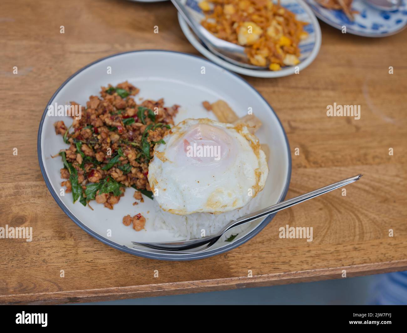 Pad Krapow MOO - Thailändisches Basilikum aus Schweinefleisch mit Reis und gebratenem Ei im Hintergrund. Thailändische Küche Gericht mit gehacktem Schweinefleisch, Basilikum, Soja und Austernsaucen. Thai Food Co Stockfoto