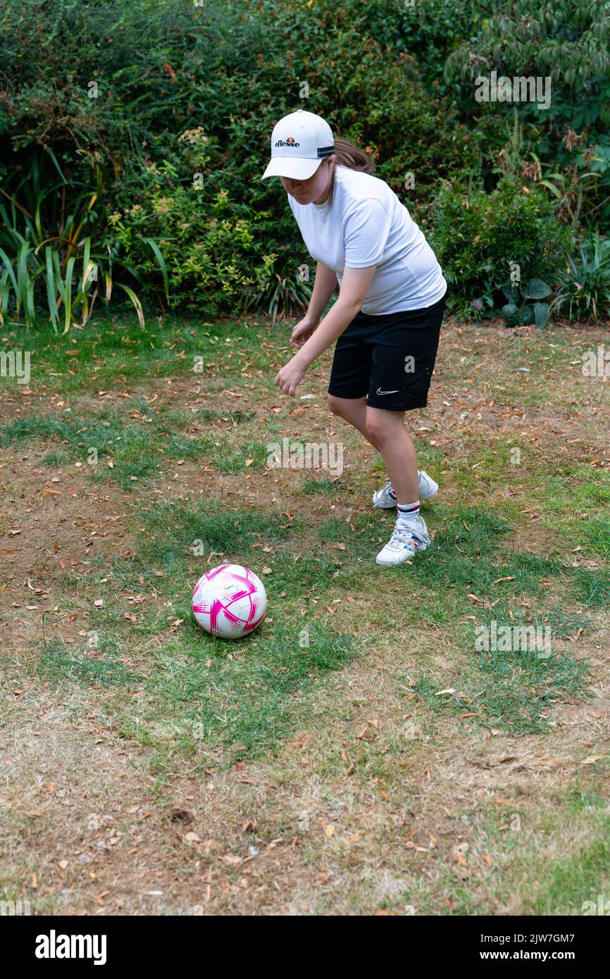 Junge Person, die im Garten Fußball spielt, mit weißer Mütze und Sportausrüstung Stockfoto
