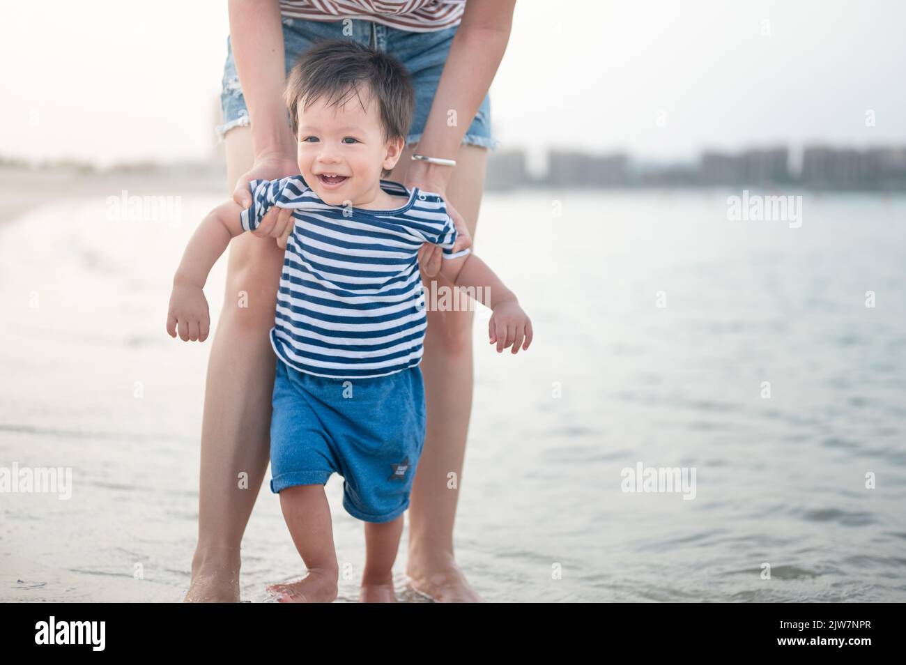 Entzückender kleiner Junge, der mit seiner Mutter seine ersten Schritte am Strand am Meer macht. Ein einjähriges Kleinkind lernt in einem Sommerurlaub, wie man zu Fuß geht Stockfoto