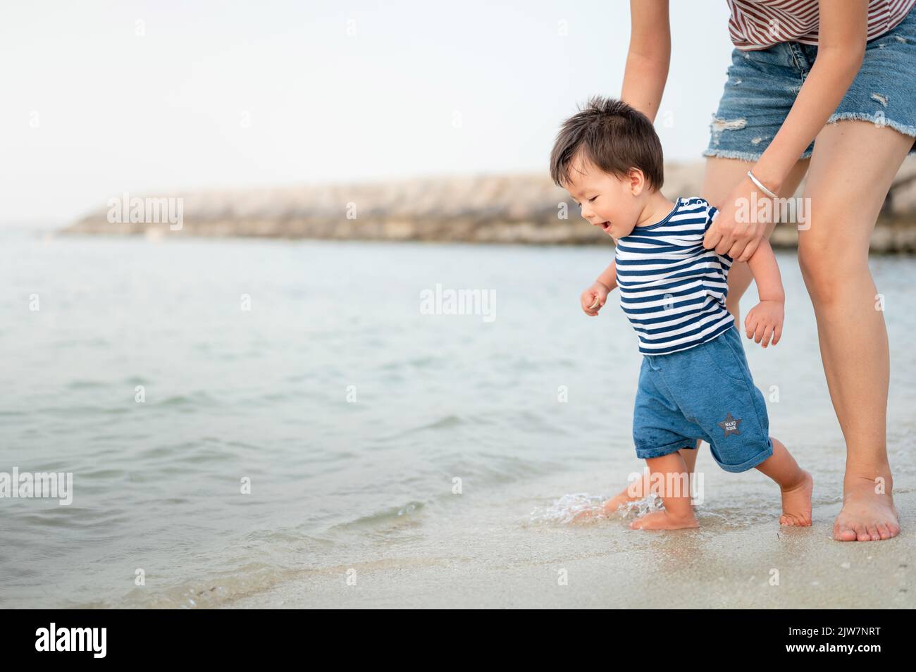 Entzückender kleiner Junge, der mit seiner Mutter seine ersten Schritte am Strand am Meer macht. Ein einjähriges Kleinkind lernt in einem Sommerurlaub, wie man zu Fuß geht. L Stockfoto