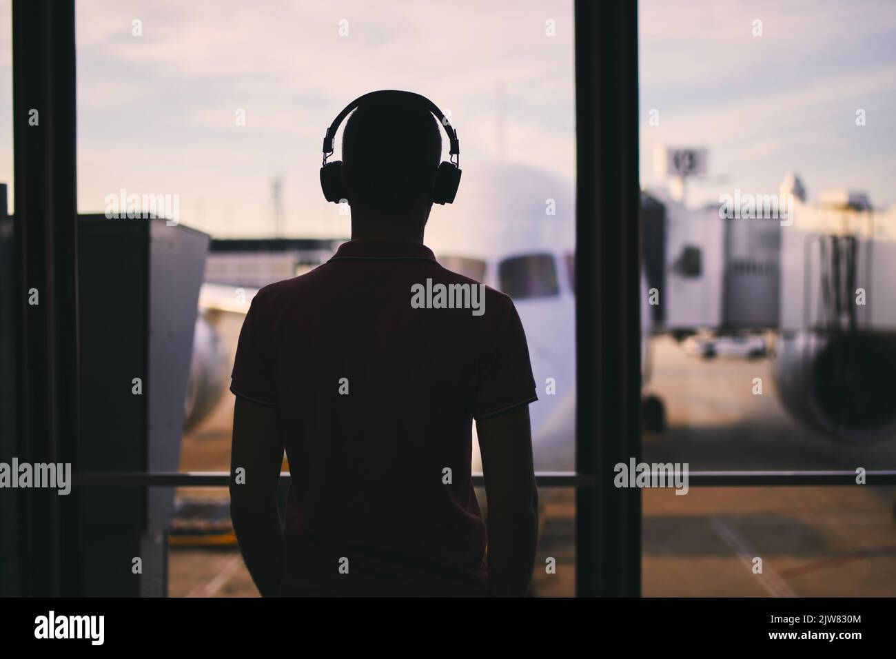 Silhouette eines Mannes mit Kopfhörern, während er auf den Flug wartet. Reisende, die vom Flughafenterminal aus schauen, bevor sie zum Flugzeug steigen. Stockfoto