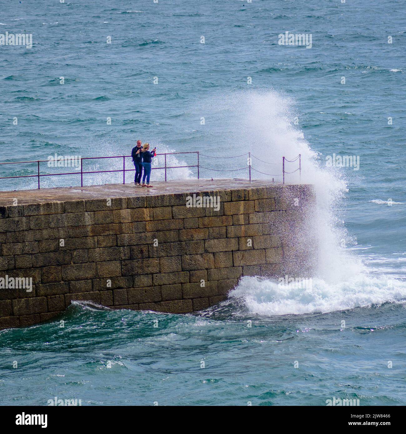 Touristen werden von großen Wellen, die über dem Porthleven Pier krachen, erwischt. Stockfoto