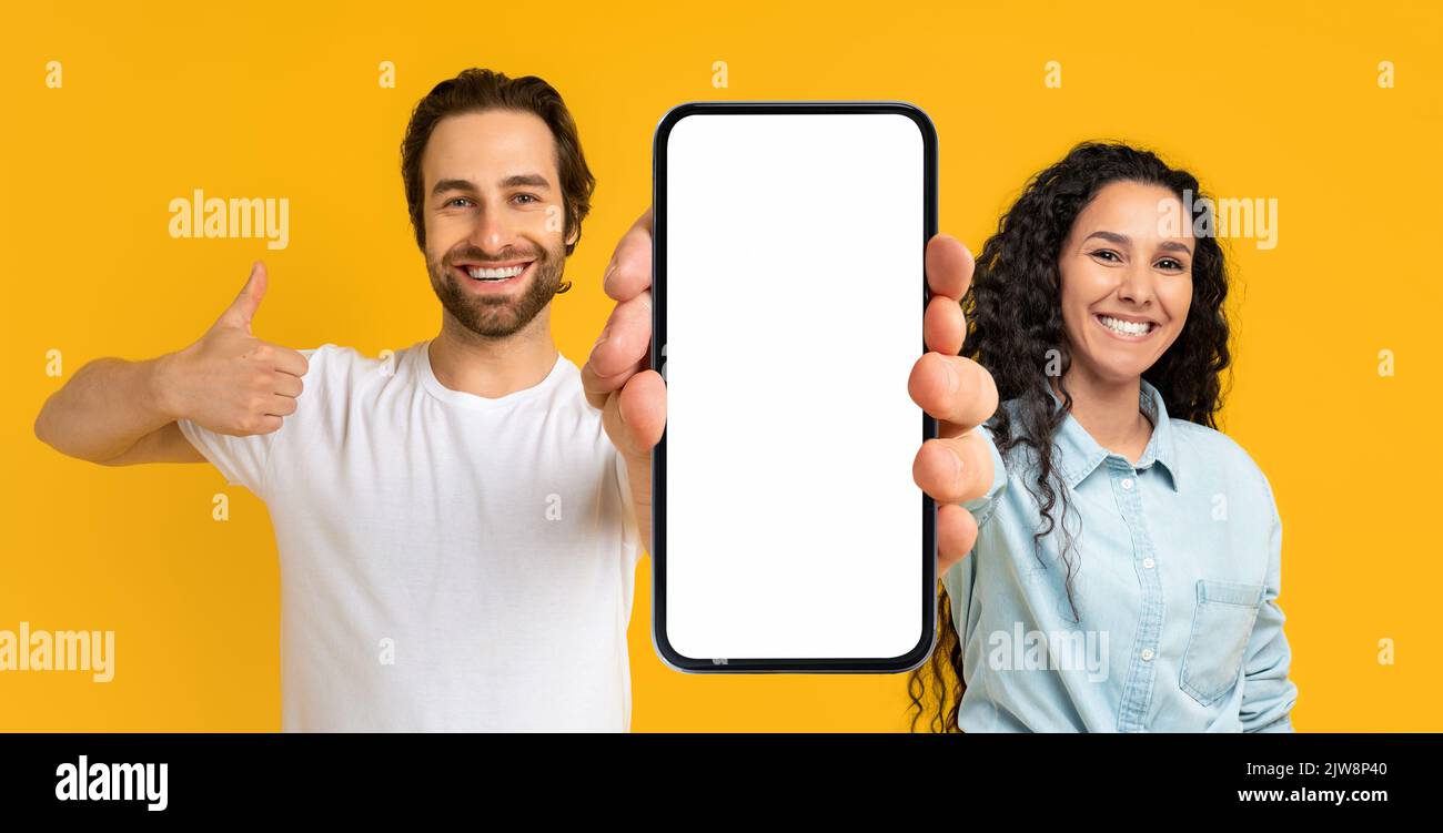 Frohes, tausendjähriges europäisches Mädchen und Mann zeigen ein großes Smartphone mit leerem Bildschirm und Daumen nach oben Stockfoto