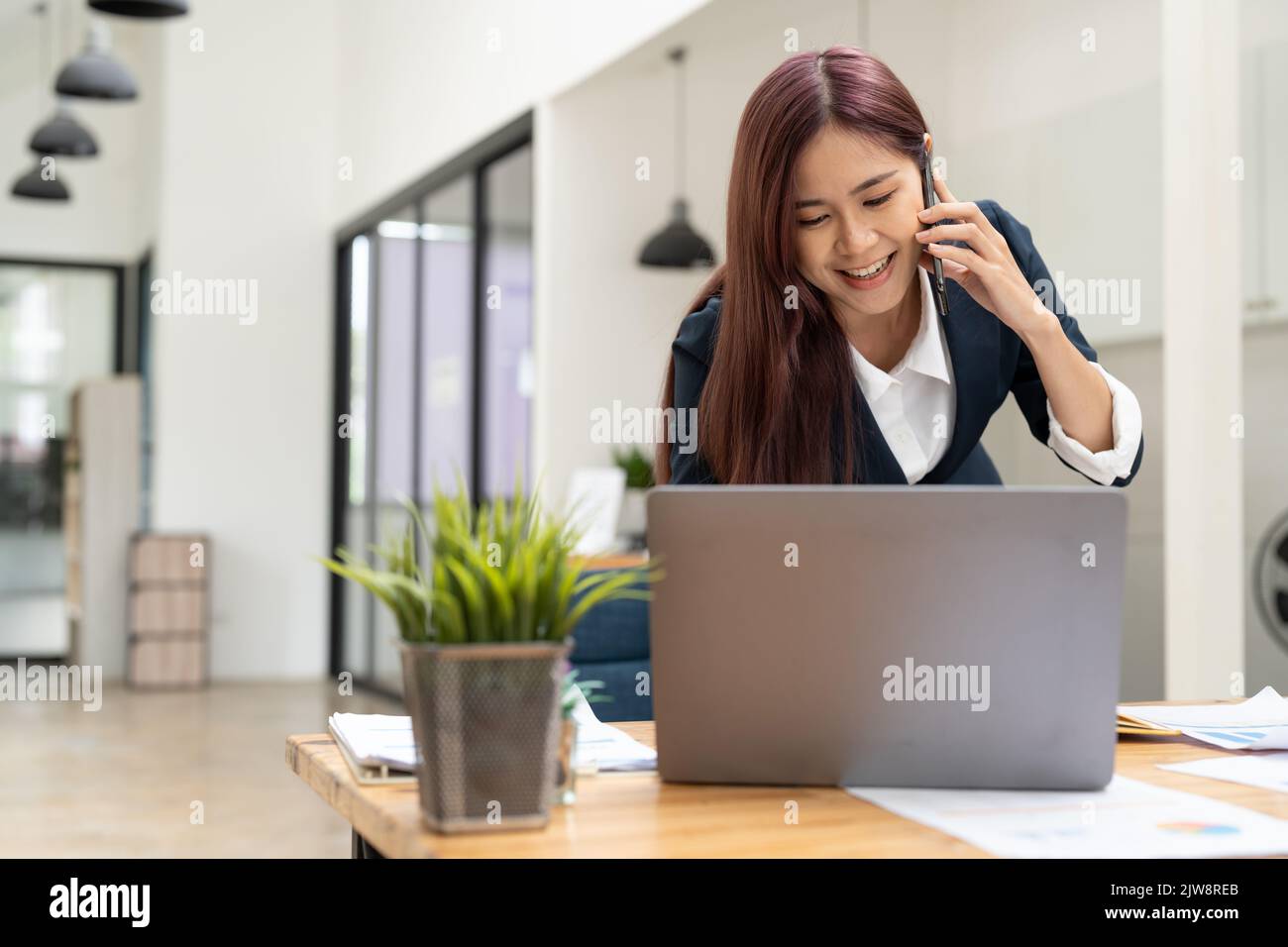 Fröhliche junge schöne Frau, die auf dem Mobiltelefon redet und mit einem Laptop lächelt, während sie an ihrem Arbeitsplatz sitzt. Copyspace. Stockfoto