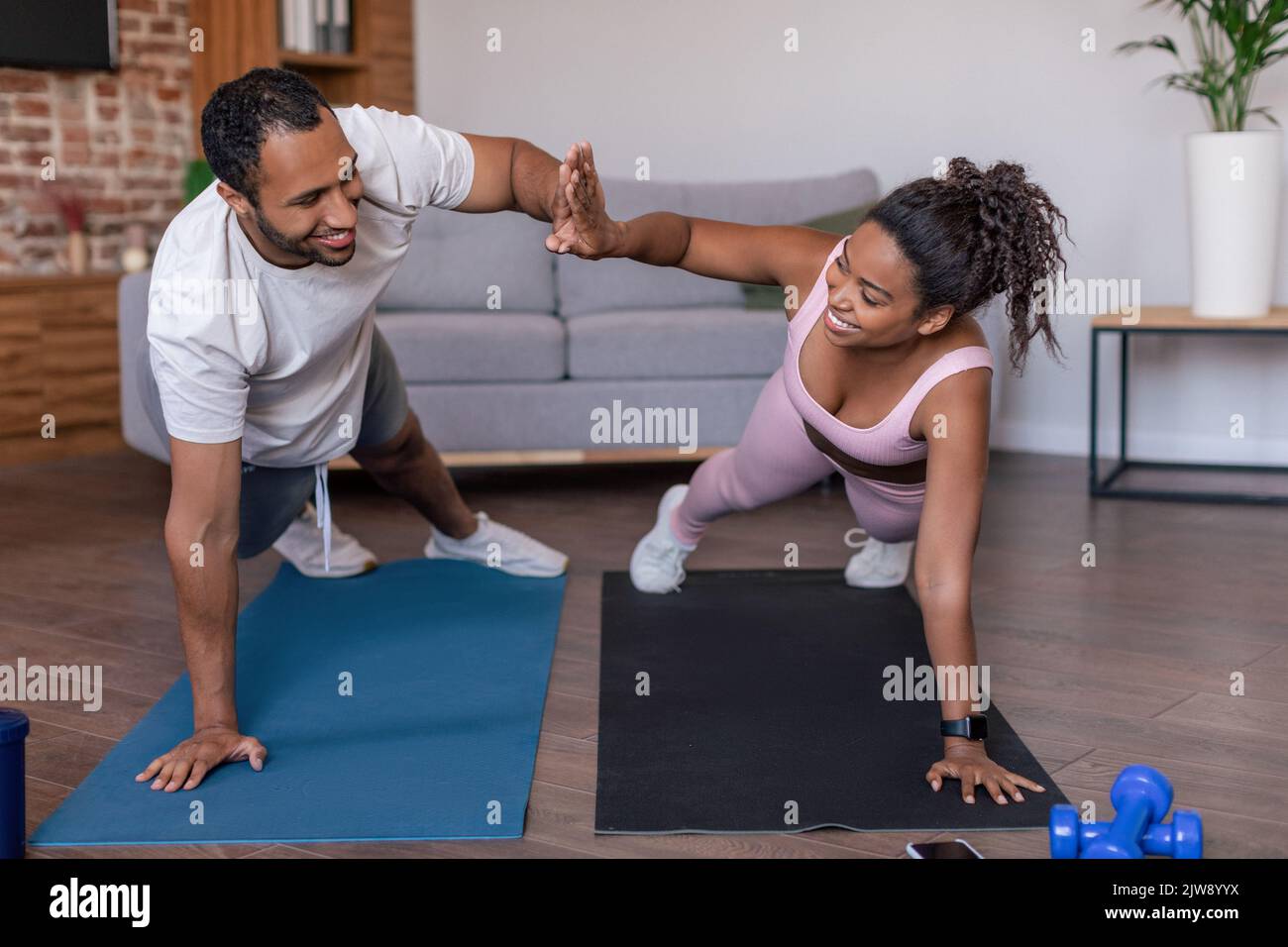 Lächelnde junge afroamerikanische Dame und Mann in Sportkleidung, die Plank-Übungen machen und hohe fünf geben Stockfoto
