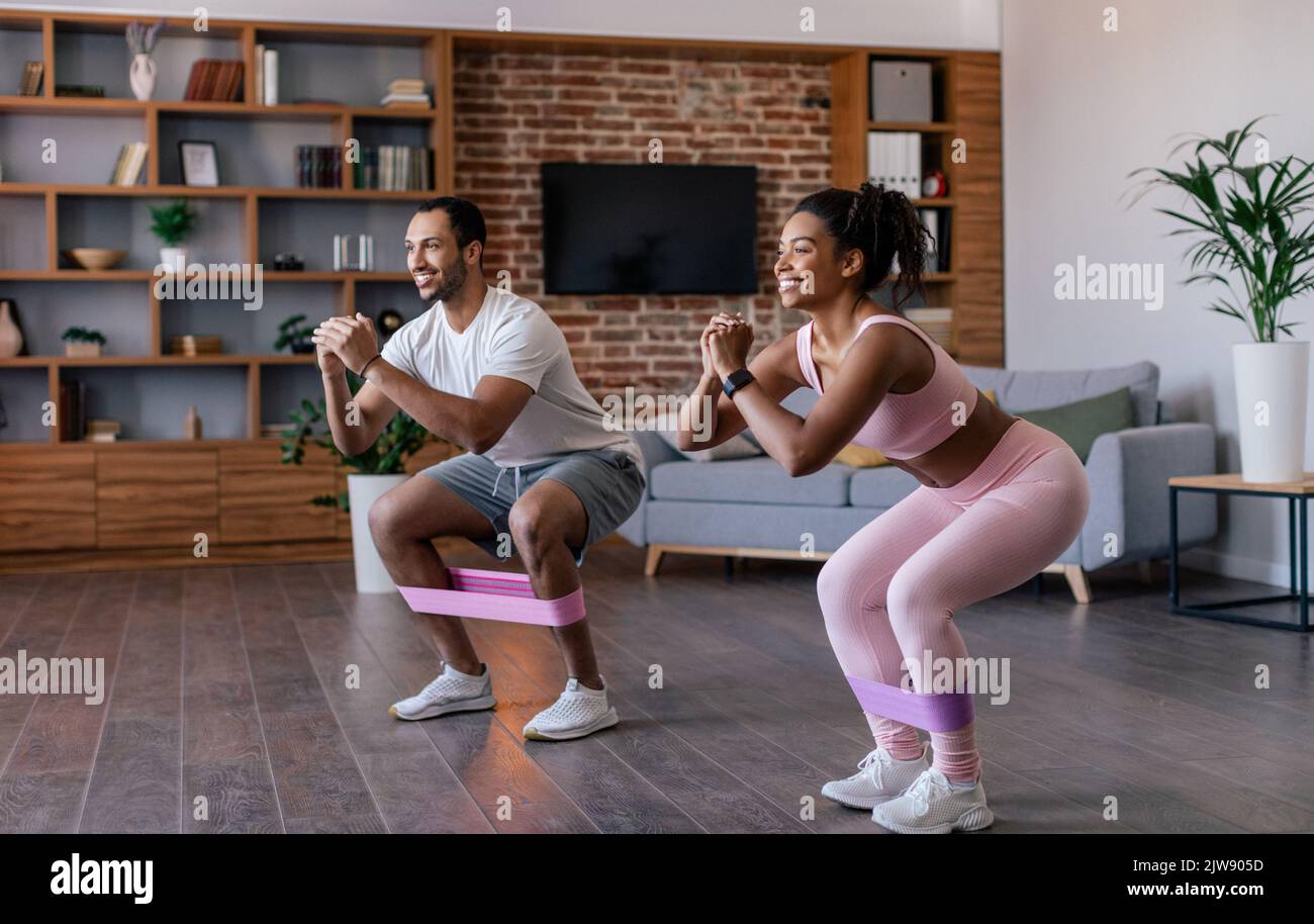 Fröhlicher junger afroamerikanischer Mann und eine junge Frau in Sportbekleidung machen Kniebeugen und Beinübungen mit Gummiband Stockfoto