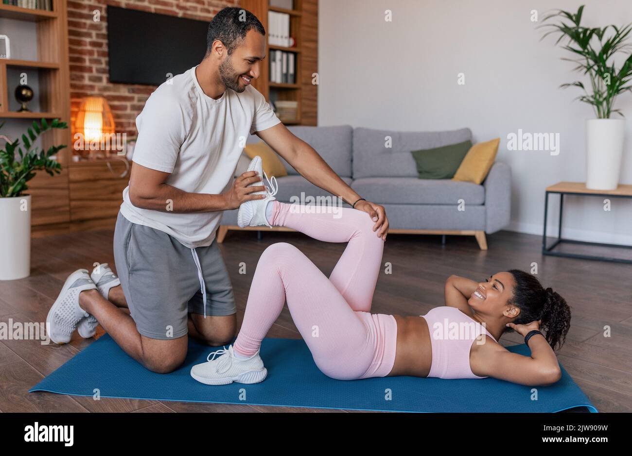 Frohes junges afroamerikanisches Männchen hilft Weibchen in Sportkleidung, Beinübungen auf Matte im Wohnzimmer zu machen Stockfoto