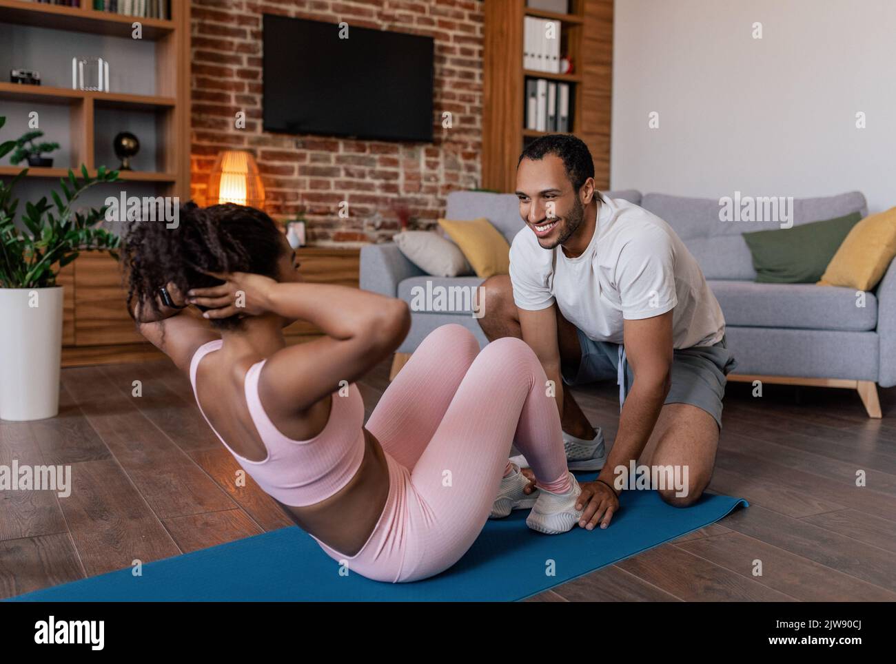 Froher junger afroamerikanischer Mann hilft der Dame in der Sportbekleidung ABS-Übungen auf der Matte zu machen, genießen Sie das Training Stockfoto