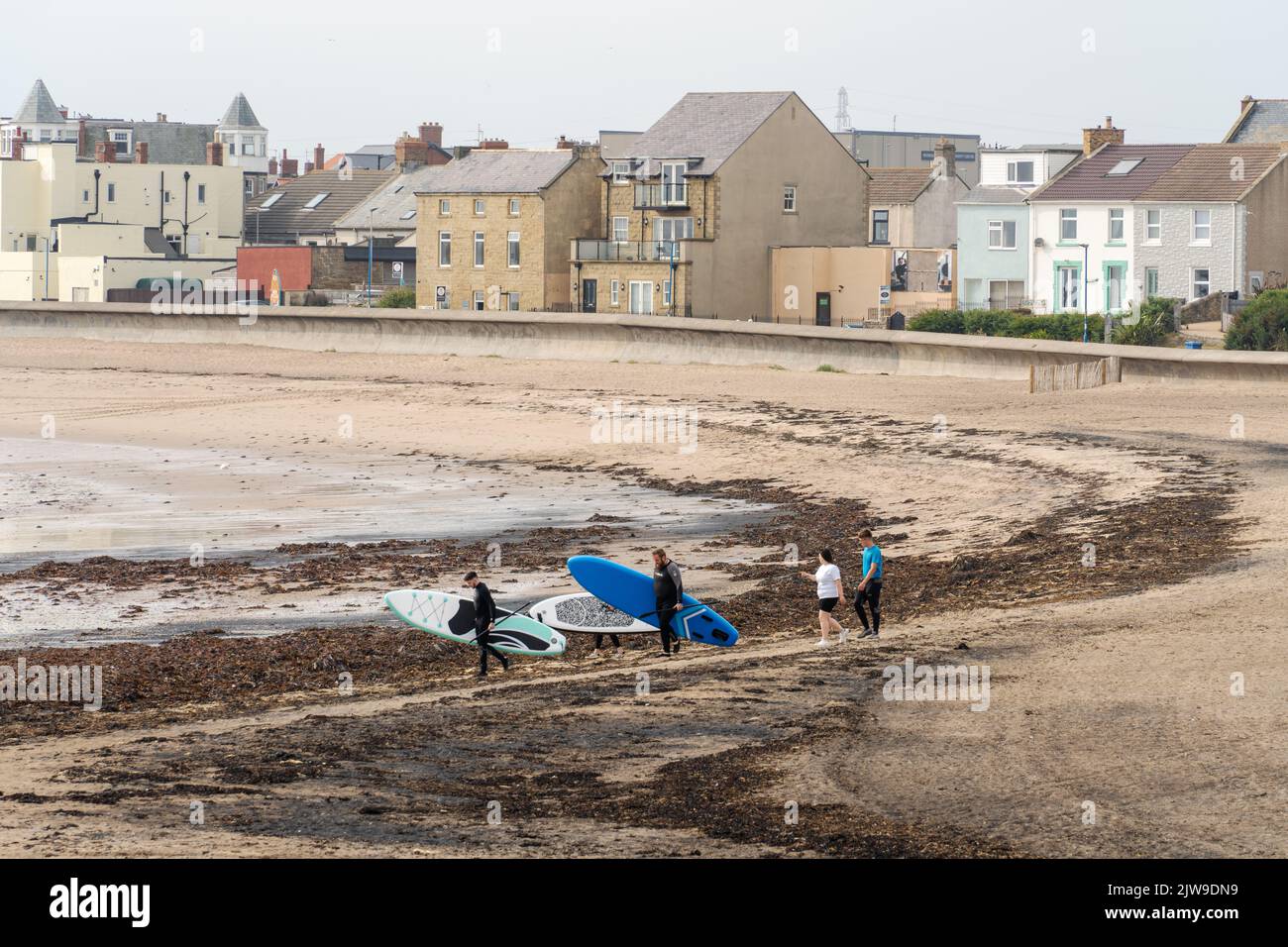 Eine Gruppe von Paddlebardern geht den Strand entlang in Richtung Meer mit ihren aufgeblasenen Brettern in Newbiggin by the Sea, Northumberland, Großbritannien. Stockfoto