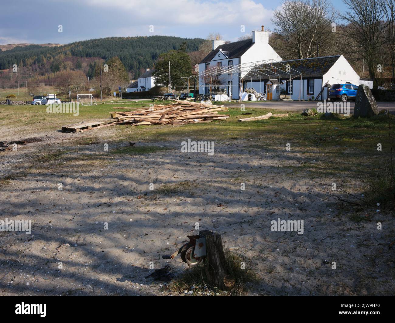 Eine rostende Winde im Vordergrund, ein Holzhaufen und ein Festzeltrahmen bedecken die Vorderansicht des Otter Ferry Inn über die Küste von Loch Fyne Stockfoto