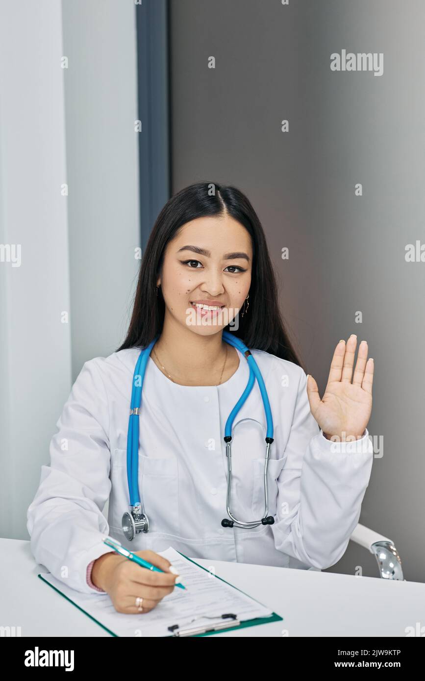 Freundliche Ärztin, die in medizinischer Uniform an seinem Arbeitsplatz sitzt und im Krankenhaus mit der Hand winkt Stockfoto