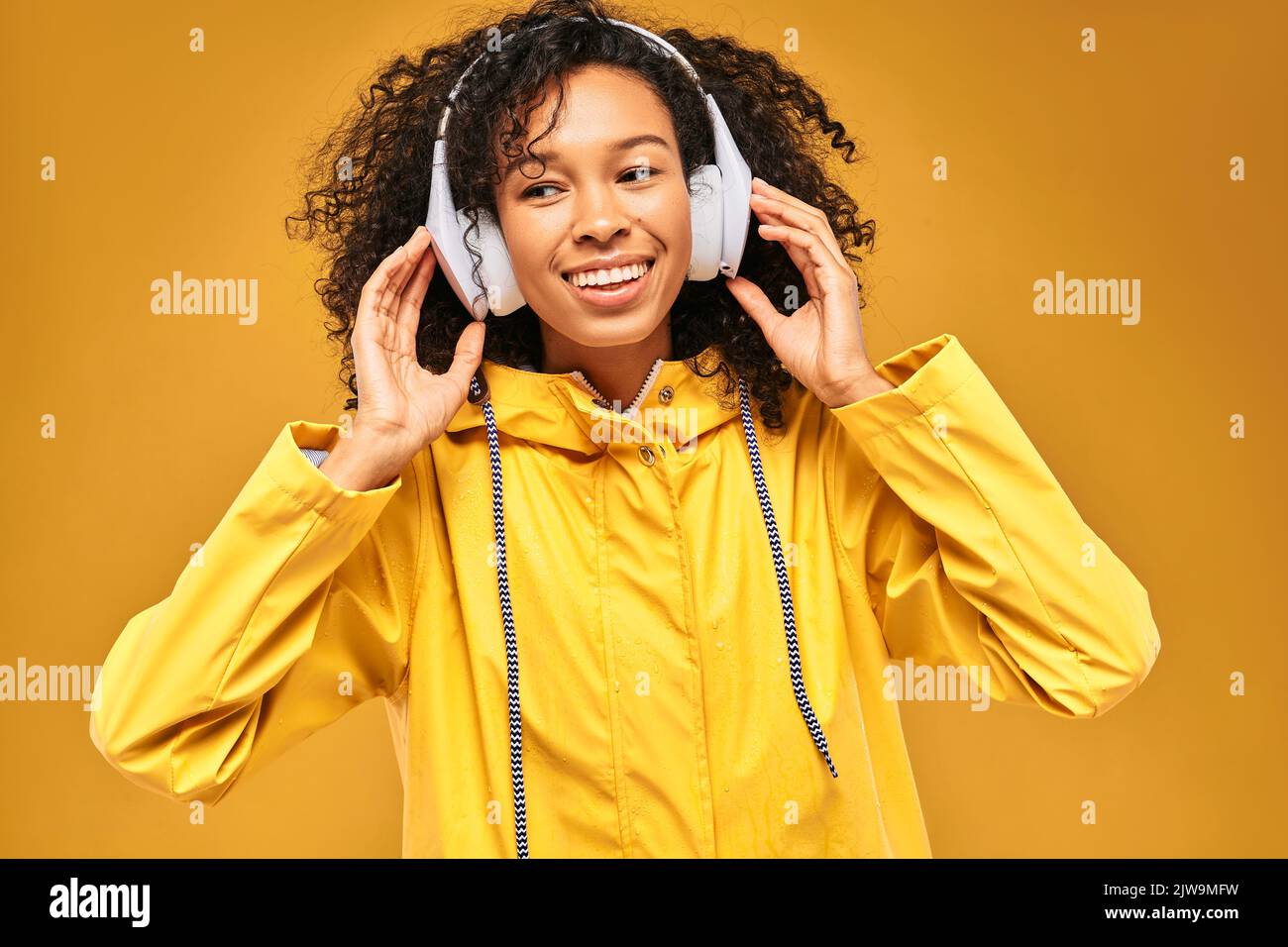 Lächelnde afroamerikanische Frau mit weißen Kopfhörern in gelbem Regenmantel mit Wassertropfen, die Musik auf gelbem Hintergrund genießen Stockfoto