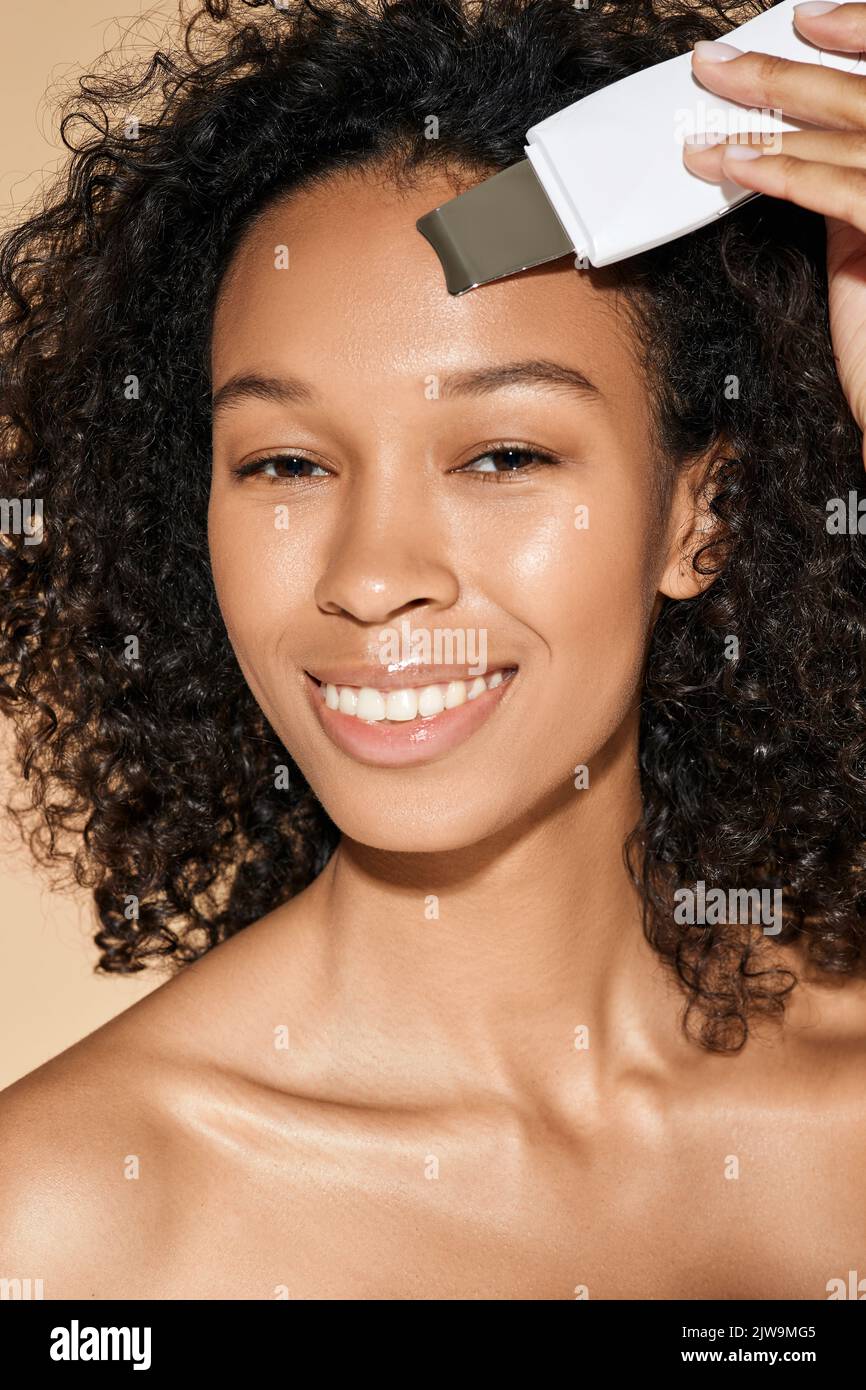 Lächelnde, lockige afroamerikanische Frau, die sich mit Ultraschallschrubber um ihre Haut kümmert, Akne entfernt und die Haut ihres Gesichts und ihrer Stirn reinigt Stockfoto