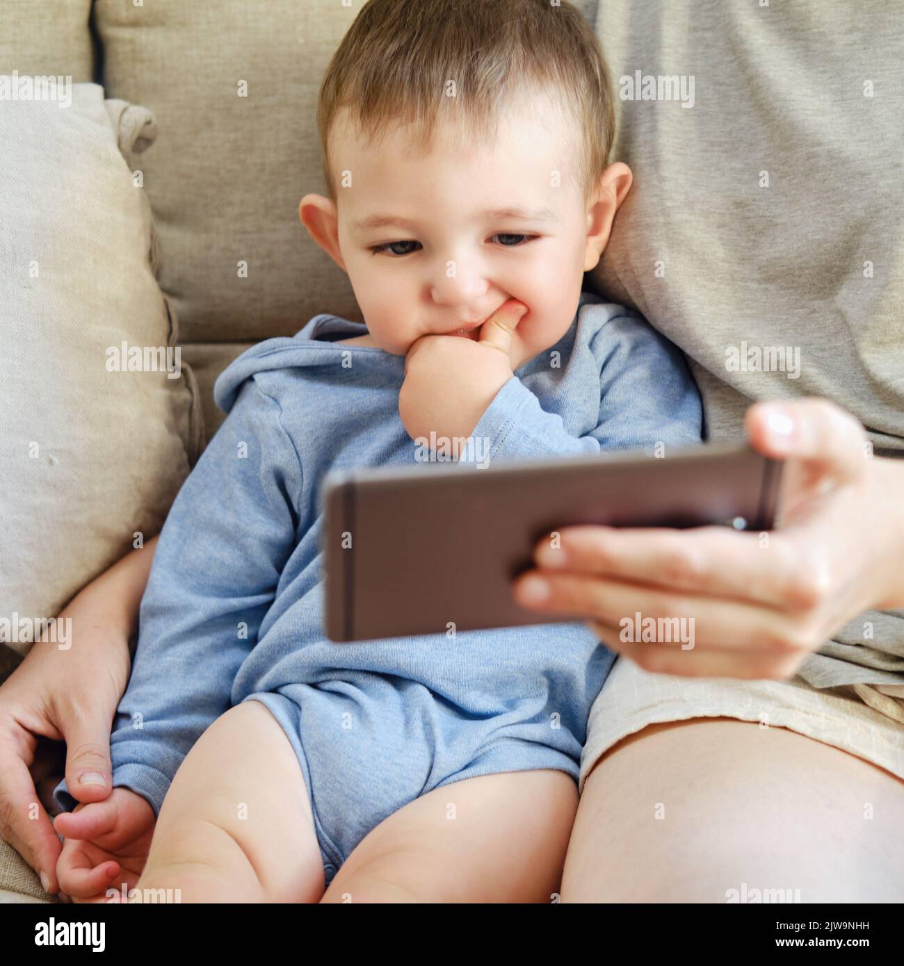 Kleinkind Baby sitzt mit seiner Mutter und schaut in das Telefon. Mutter und Kind mit einem Smartphone in den Händen auf dem Sofa Stockfoto