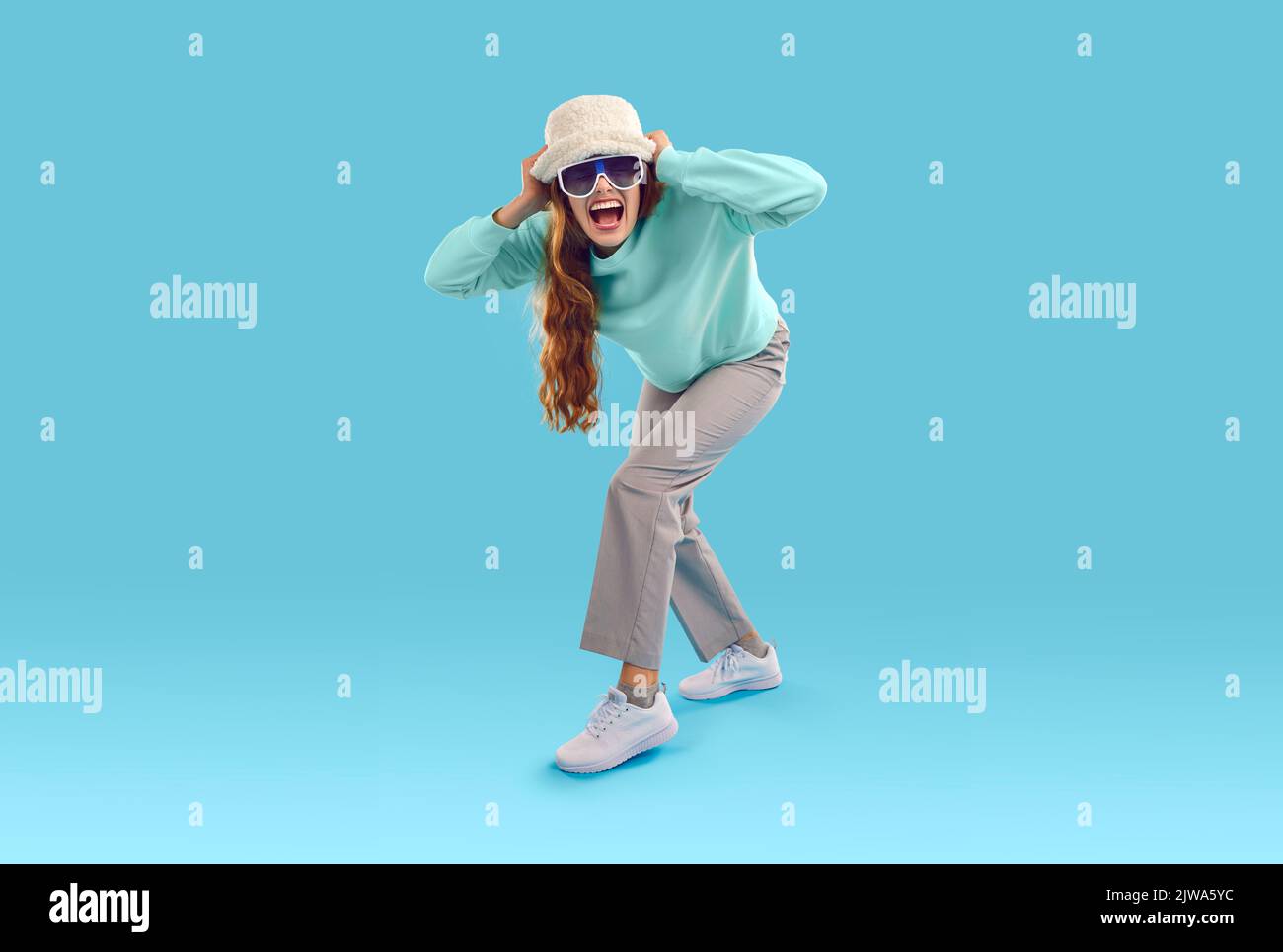 Lustige, aufgeregte Frau mit Hut und Sonnenbrille, die auf blauem Studiohintergrund steht und schreit Stockfoto