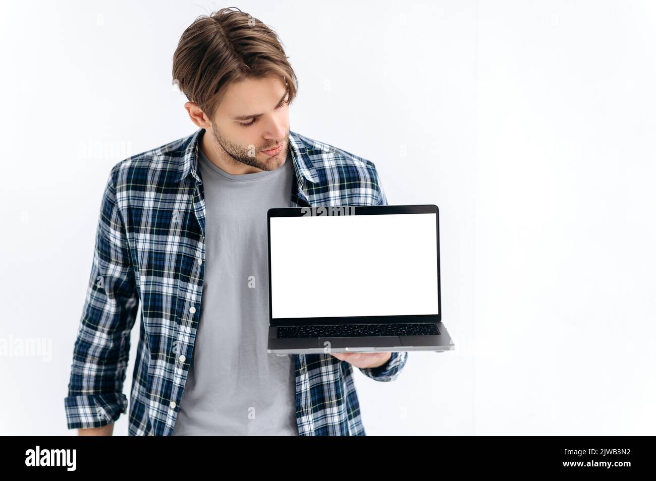 Ein moderner, gutaussehender, selbstbewusster Kaukasier, der auf einem weißen, isolierten Hintergrund steht, hält einen offenen Laptop, zeigt einen leeren weißen Bildschirm für Werbung oder Präsentation und sieht ihn an Stockfoto
