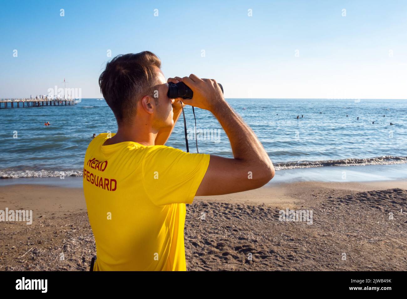 Rettungsschwimmer am Strand mit Blick durch ein Fernglas. Rückansicht des hübschen brünette männlichen Rettungsschwimmers mit gelbem Hemd am Strand im Sommer. Stockfoto