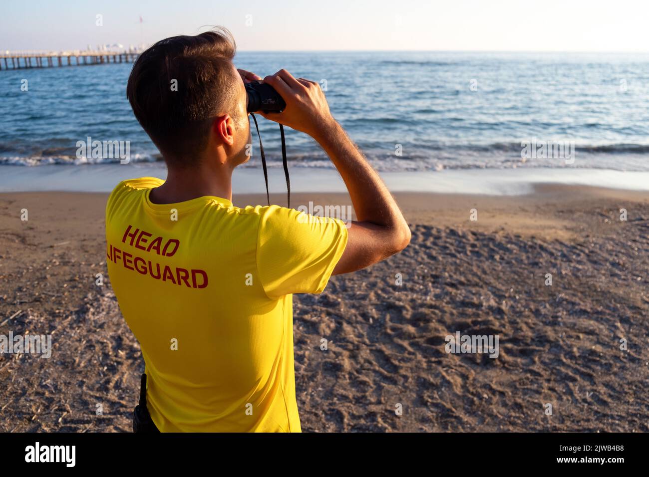 Rettungsschwimmer am Strand mit Blick durch ein Fernglas. Rückansicht des hübschen brünette männlichen Rettungsschwimmers mit gelbem Hemd am Strand im Sommer. Stockfoto