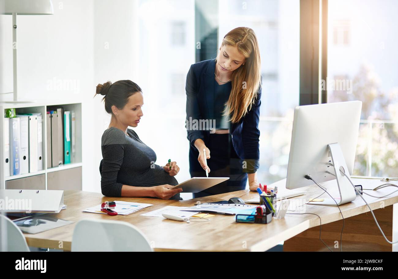 Sehen Sie selbst: Eine Schwangere Geschäftsfrau und ein Kollege, die in ihrem Büro ein digitales Tablet benutzen. Stockfoto