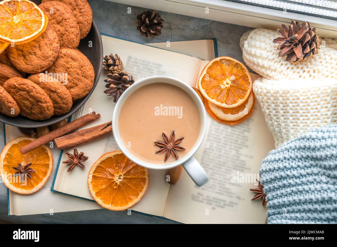 Eine große Tasse Kaffee und ein Buch am Fenster, mehrere Strickdecken und Scheiben getrockneter Orangen erinnern an den Herbst, eine gemütliche Zeit und einen Urlaub zu Hause. Flach liegend Stockfoto