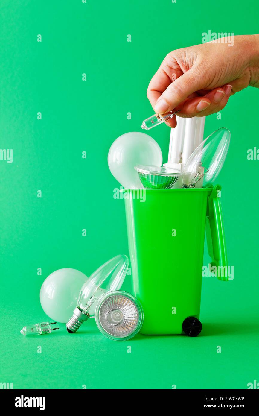 Grünes Wohnkonzept: Diverse alte ineffiziente Glühbirnen in und um einen grünen Recyclingbehälter mit einer Hand, die eine weitere Halogenlampe wegwirft. Stockfoto