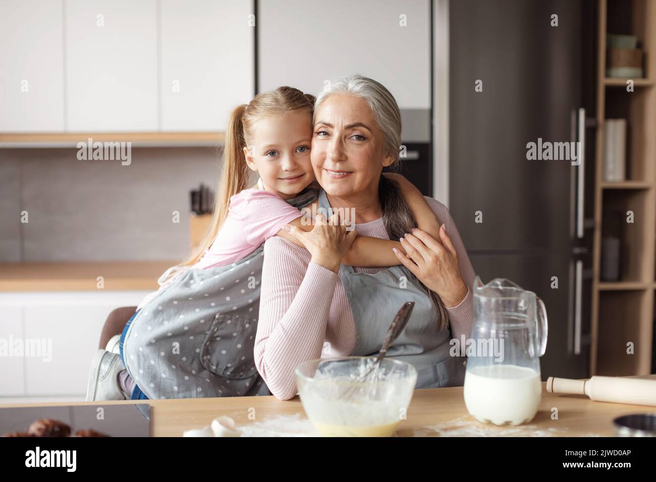 Lächelndes kleines Mädchen umarmt die alte Frau in der Schürze, die Teig in der Küche macht Stockfoto