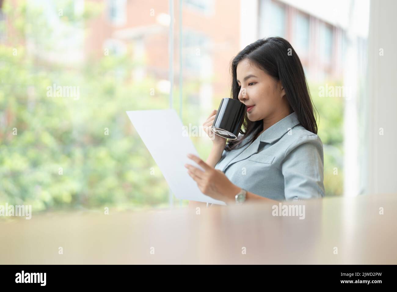 Schöne asiatische Geschäftsfrau in Eleganz Anzug sitzen am Schreibtisch und hält eine Kaffeetasse. Unternehmerin, Geschäftsbesitzerin. Stockfoto