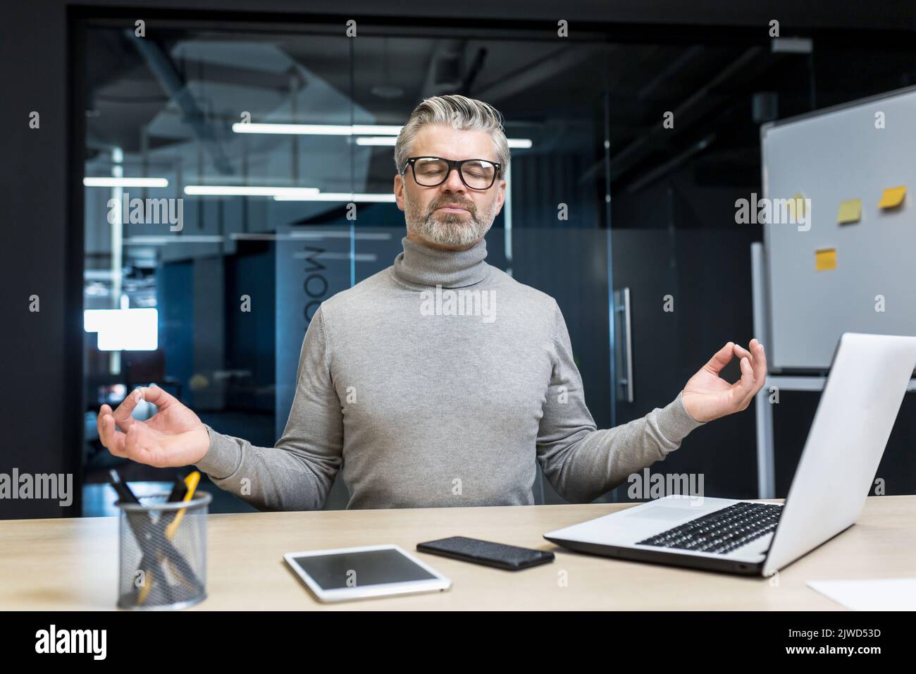 Ein grauhaariger Geschäftsmann, der im Bürogebäude meditiert, sitzt auf einem Stuhl am Schreibtisch, ein reifer Mann trägt eine Brille mit geschlossenen Augen in Lotusposition, ein Chef arbeitet am Arbeitsplatz mit einem Laptop. Stockfoto