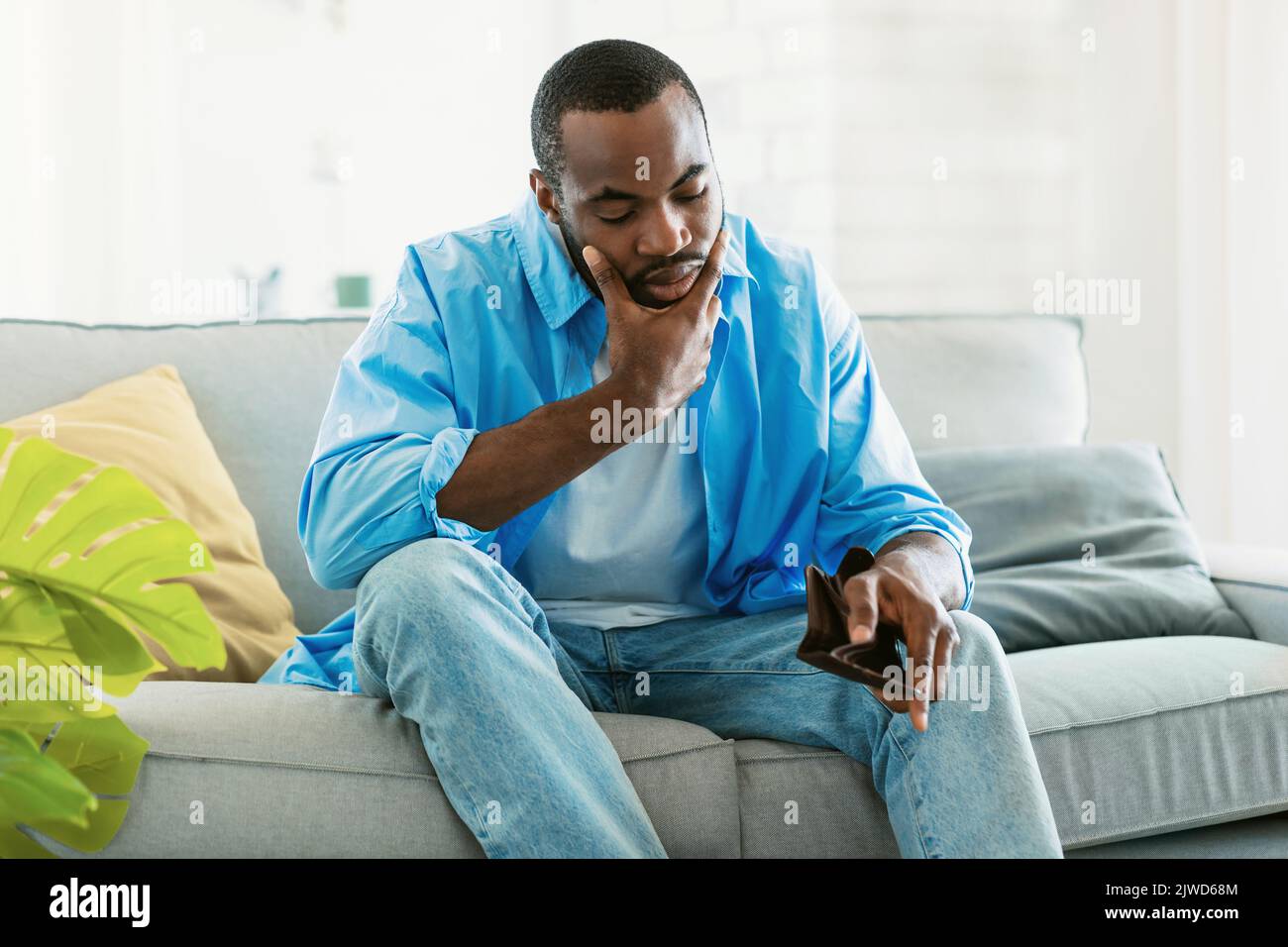 Ich bin pleite. Trauriger afroamerikanischer Mann pleite, zeigte seine leere Brieftasche und schaute sie an, saß zu Hause auf dem Sofa Stockfoto