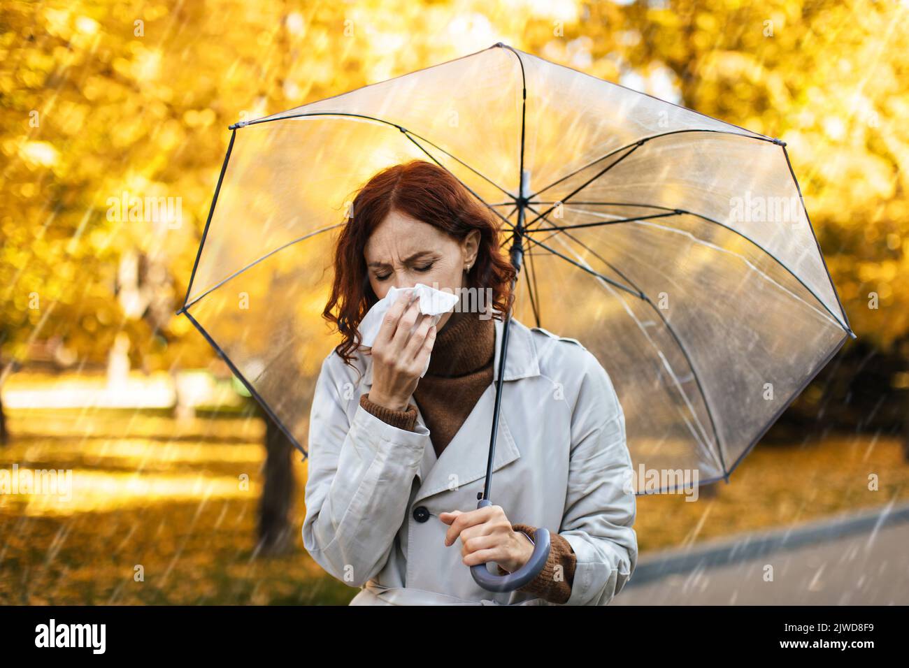 Unglückliche, tausendjährige europäische Frau im Regenmantel mit Regenschirm bläst ihre Nase auf Serviette, fühlt sich im Park schlecht Stockfoto