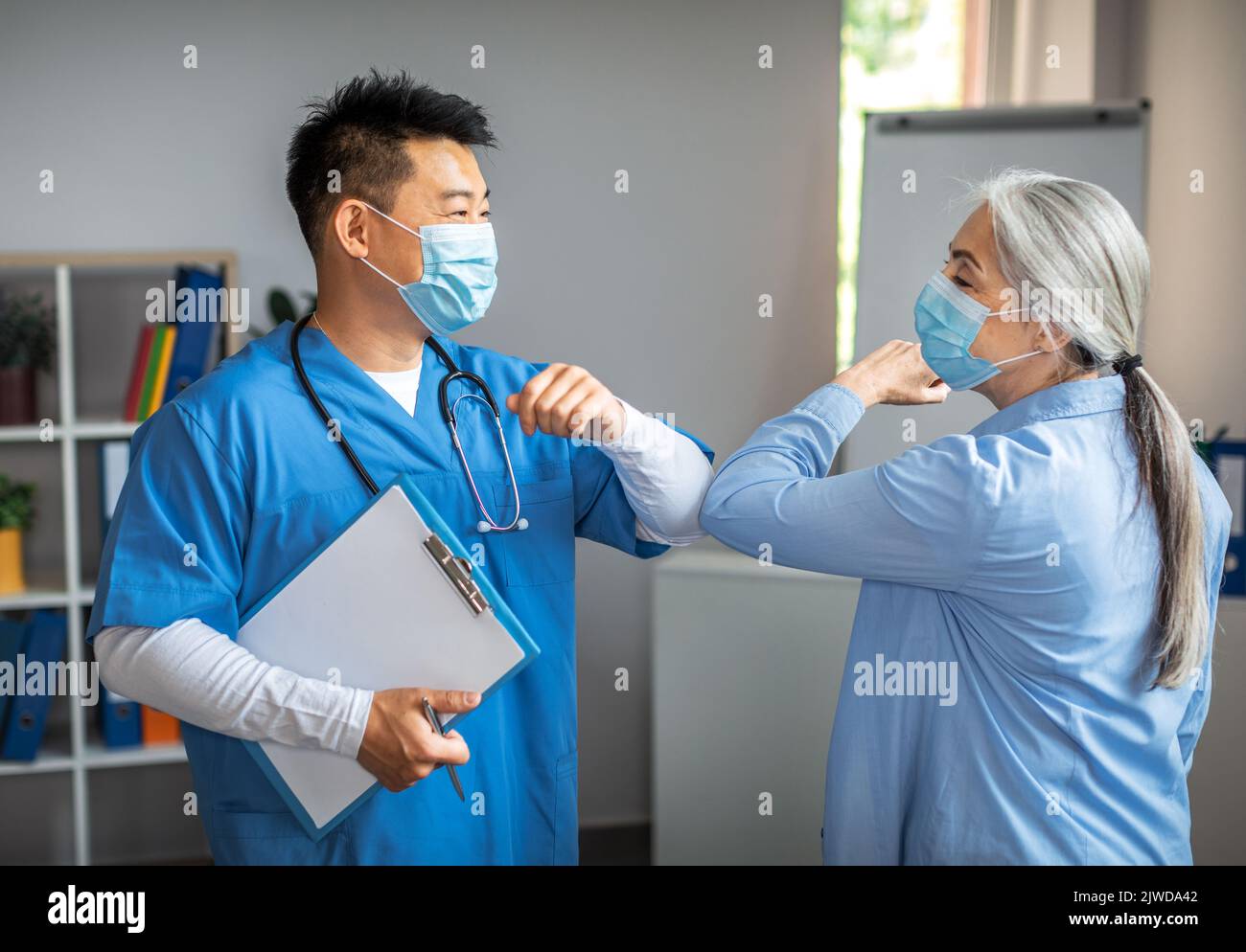 Lächelnder erwachsener chinesischer Mann, Arzt in Schutzmaske, begrüßt die alte Patientin in der Klinik mit Ellbogen Stockfoto