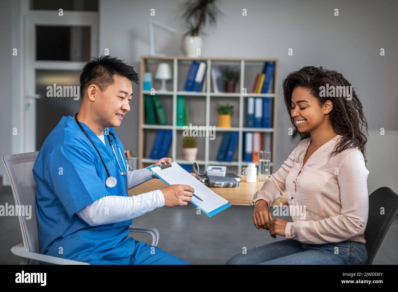 Glücklicher erwachsener chinesischer Arzt, der in der Klinik einen Vertrag mit einer jungen afroamerikanischen Patientin zeigt Stockfoto