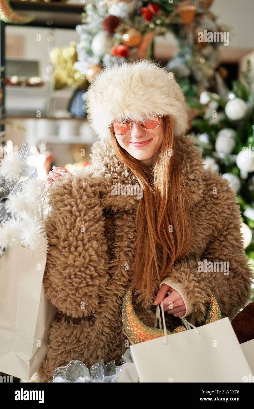 Attraktive kaukasische Mädchen in warmer Kleidung und Sonnenbrille mit Einkaufstaschen voller Weihnachtsdekoration im Laden oder Supermarkt. Neujahrseinkäufe oder Konsumdenken. Hochwertiges vertikales Bild Stockfoto