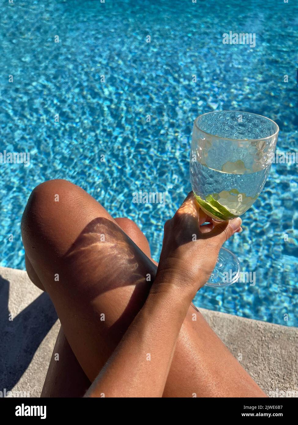 Frau, die mit gekreuzten Beinen am Rand eines Schwimmbeckens sitzt und ein Glas Kalkwasser hält Stockfoto