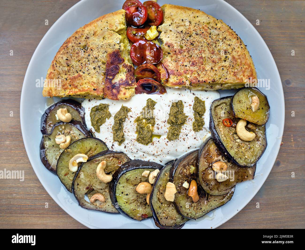 Überzogene Mahlzeit von oben mit spanischer Tortilla und gebratenen Auberginen-Scheiben mit Nüssen und grobem Pfeffer. Serviert mit Joghurt mit Kräutern und kleiner Kirsch-Toma Stockfoto