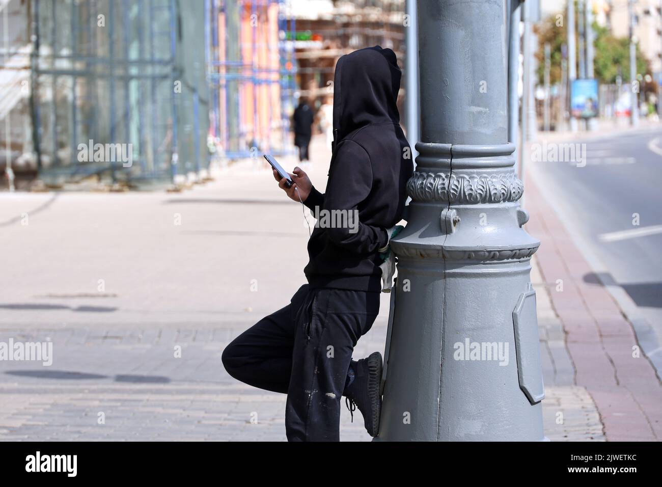 Kerl in schwarzem Kapuzenpullover mit Smartphone an einen Pfosten gelehnt. Schläger auf der Stadtstraße Stockfoto