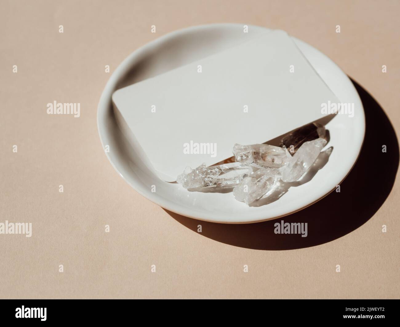 Weiße Visitenkartenvorlage auf weißem Teller mit hellen Kristallen auf beigem Hintergrund. Design-Element für Hochzeitseinladung, danke Stockfoto