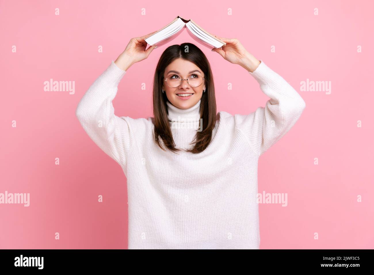 Lächelndes Mädchen, das den Kopf mit einem Buch bedeckt und mit klugen Augen weit weg sieht, träumt von einer zukünftigen Karriere und trägt einen weißen Pullover im lässigen Stil. Innenaufnahme des Studios isoliert auf rosa Hintergrund. Stockfoto