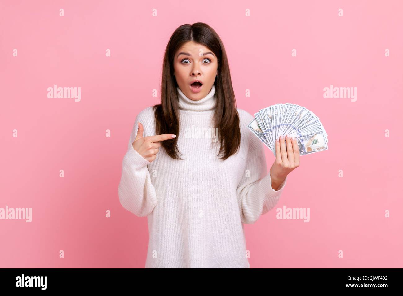 Schockiert lächelnde Frau zeigt auf Fan von Dollar-Banknoten, schaut überrascht auf die Kamera, trägt weißen Pullover im lässigen Stil. Innenaufnahme des Studios isoliert auf rosa Hintergrund. Stockfoto